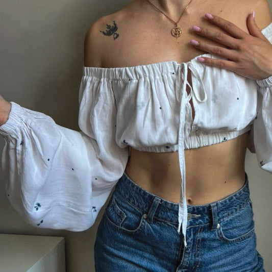 A woman wearing a white off-the-shoulder crop top with long balloon sleeves and small black embroidered details, paired with blue jeans.
