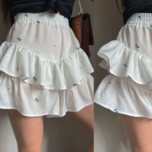 White tiered ruffle mini skirt with small blue floral embroidery, shown on a model from the side and back.