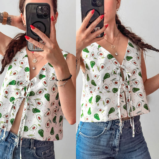 A split-screen mirror selfie showing a woman wearing a white sleeveless tie-front top with an avocado print and blue jeans. The top features a repeating pattern of green avocados and red/pink seed details on a white background.