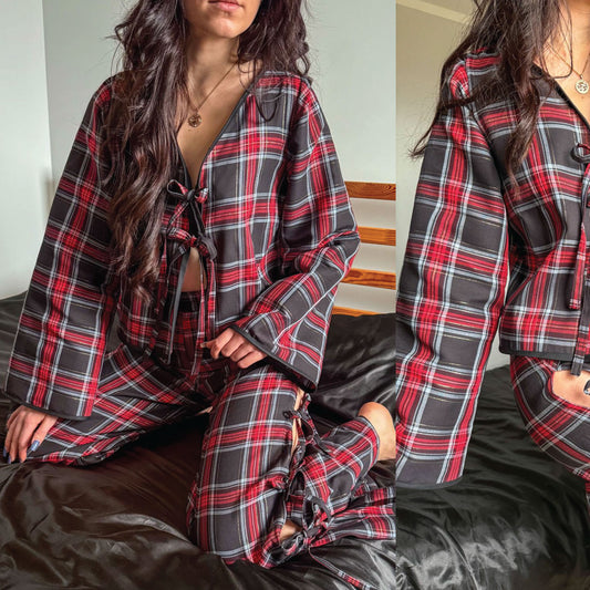 A woman wearing a matching plaid pajama set with a long-sleeve button-front top and tie-front pants, seated on a bed. The fabric has a red, black, and gray plaid pattern.