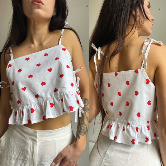 A woman modeling a white spaghetti-strap crop top with a ruffled hem and small red heart print, shown from both the front and back against a plain indoor background.