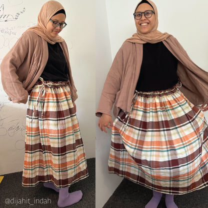 A side-by-side comparison of a woman modeling a long plaid maxi skirt, styled with a black top, a taupe cardigan, a beige hijab, and purple socks. She is standing indoors against a white wall with some faint drawings on it.