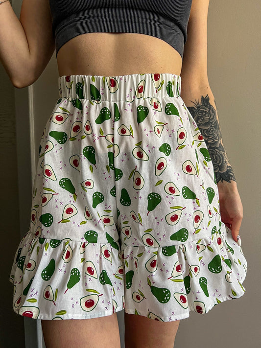 A close-up of a person wearing high-waisted white shorts with an avocado print, featuring a wide elastic waistband and a gathered ruffle at the bottom hem. The person has a tattoo on their forearm and is posed against a plain neutral wall.