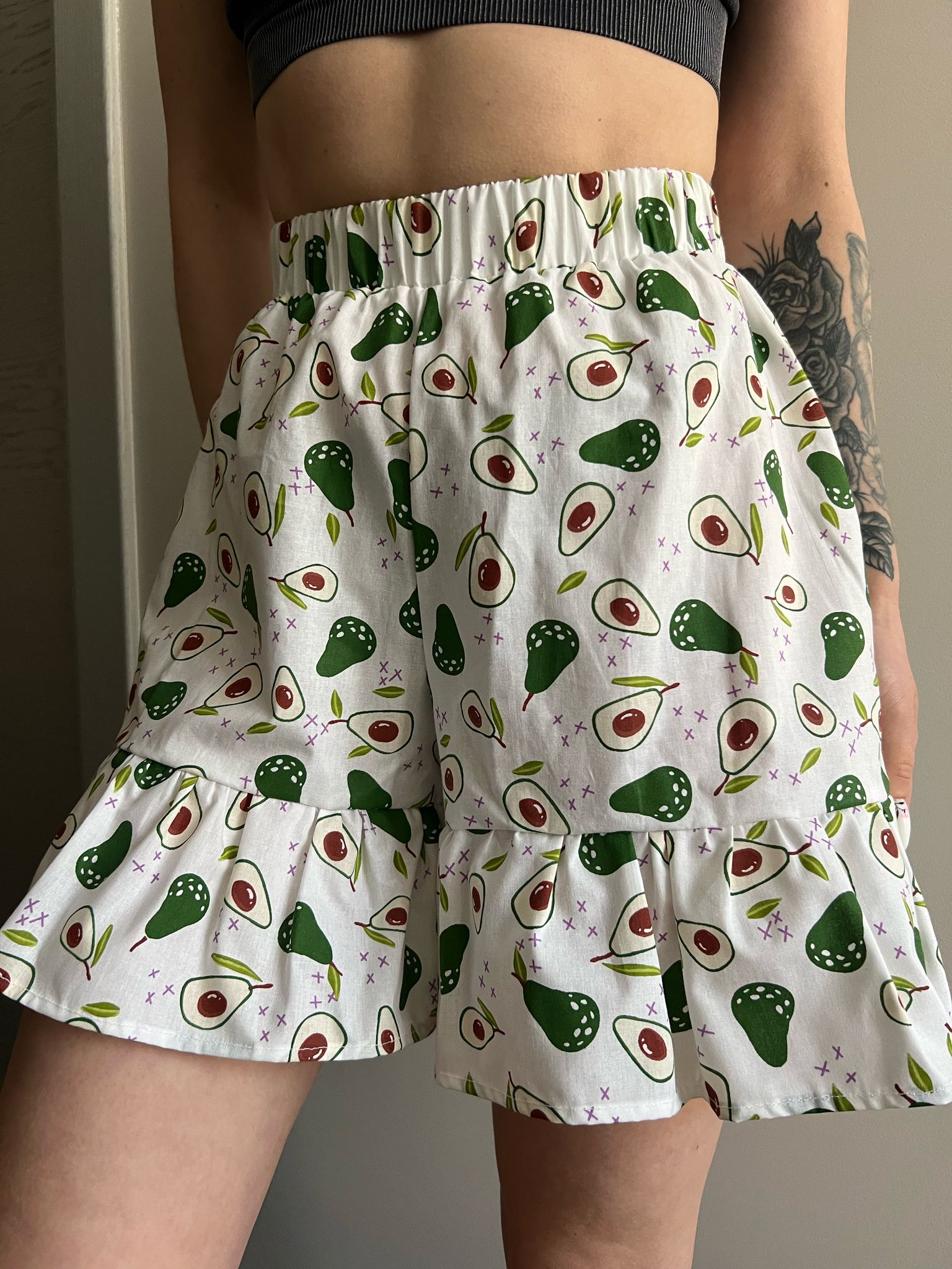 A close-up of a person wearing high-waisted white Bloom Shorts with a green and red avocado print. The shorts feature a wide elastic waistband and a gathered ruffle at the bottom hem.
