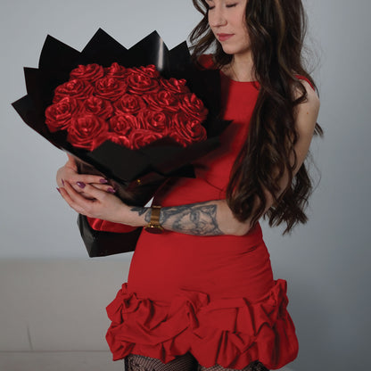 A woman in a red dress holding a large bouquet of red roses wrapped in black paper, posed against a plain light gray background.