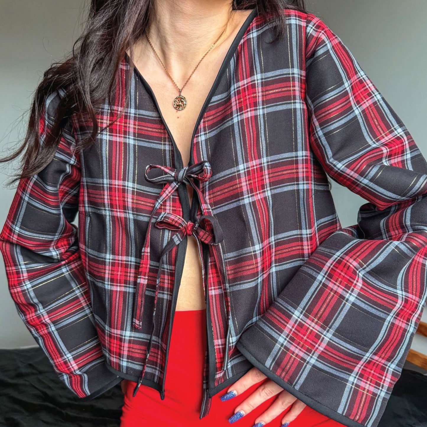 A close-up of a person wearing a long-sleeve plaid wrap-style top with a tie-front closure. The garment has a black base with red, white, and thin gray plaid lines.