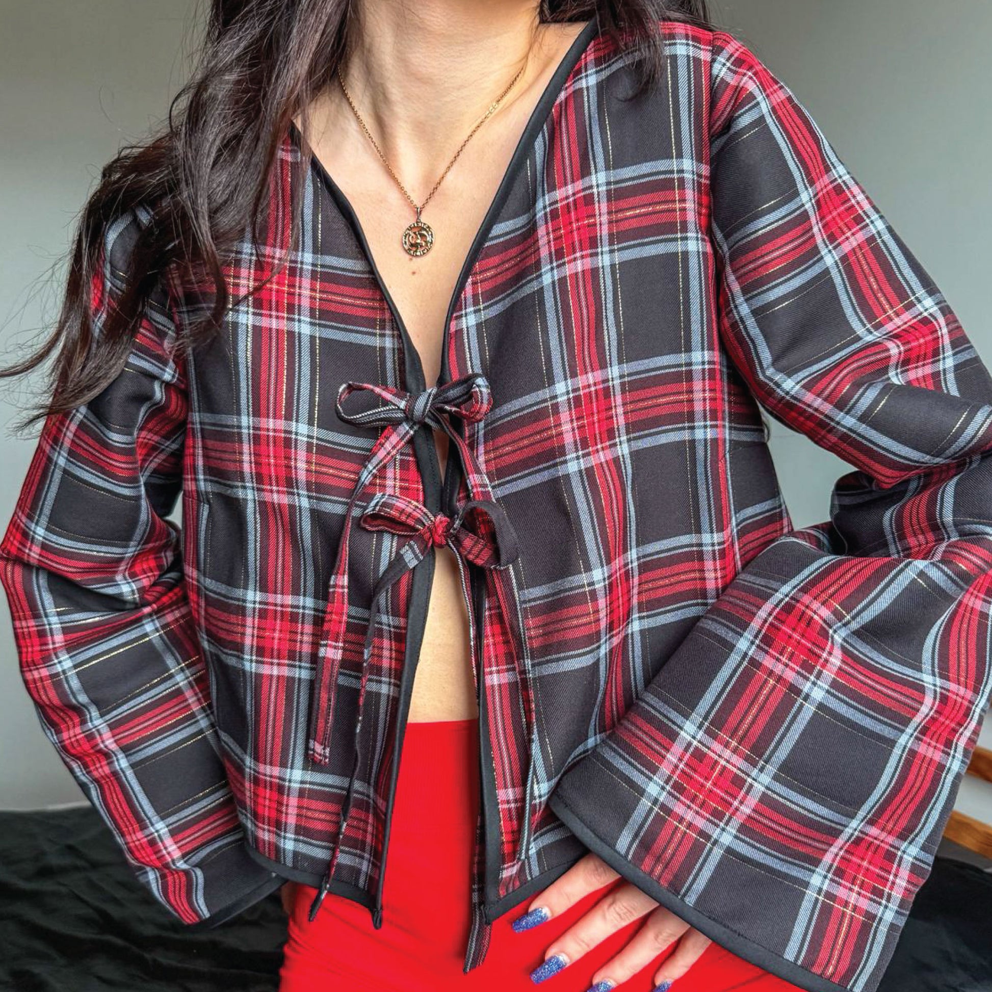 A close-up of a person wearing a long-sleeve plaid wrap-style top with a tie-front closure. The garment has a black base with red, white, and thin gray plaid lines.