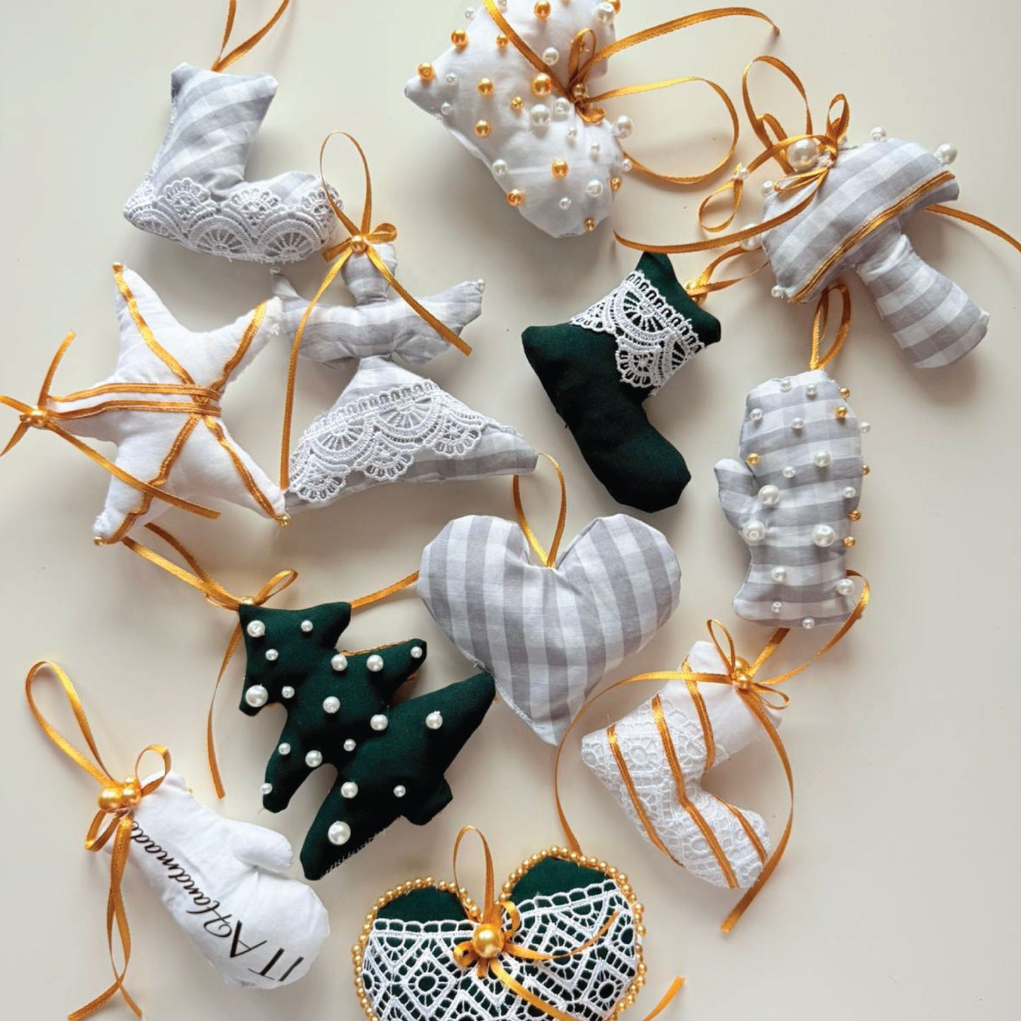 A collection of handmade Christmas fabric ornaments shaped like socks, hearts, stars, mittens, and Christmas trees, arranged on a light beige background. The ornaments feature white and gray gingham patterns, dark green velvet, white lace, gold ribbons, and small pearl embellishments.