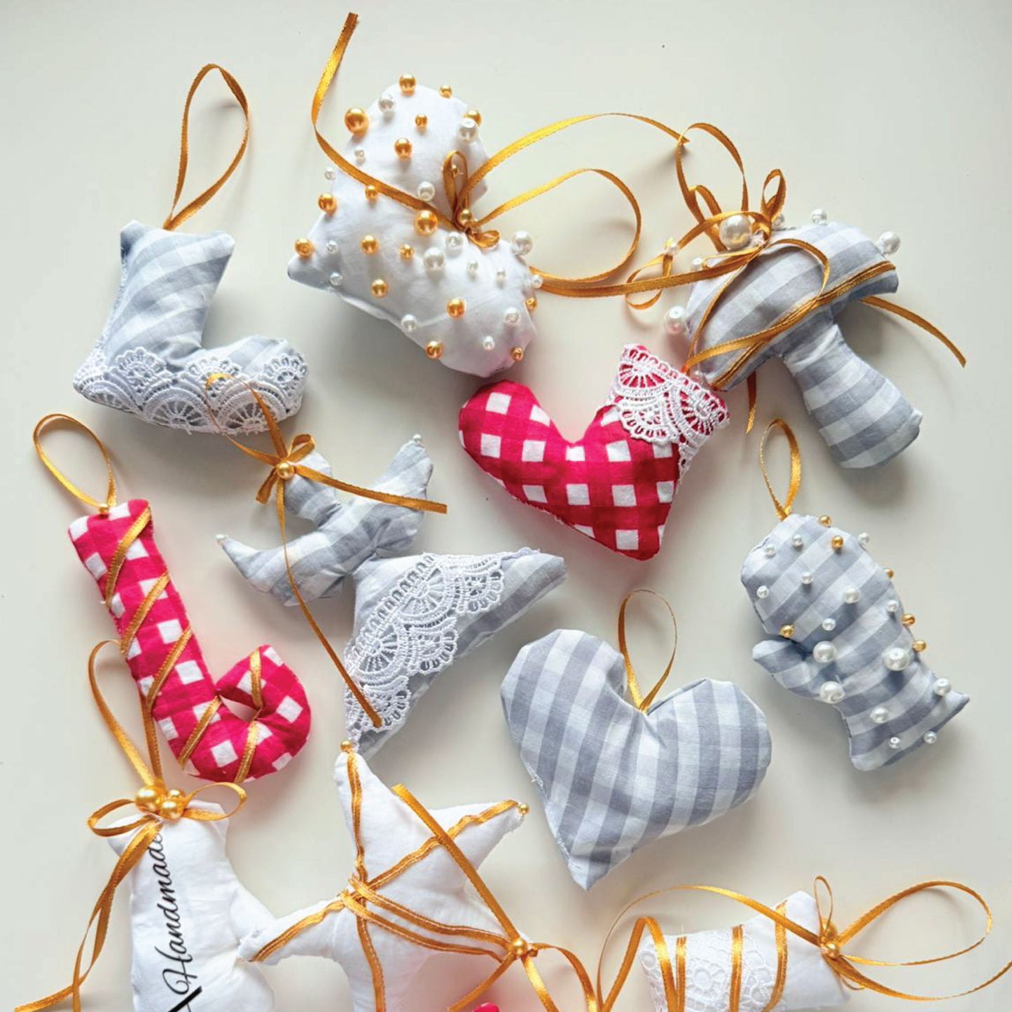A collection of handmade fabric Christmas ornaments arranged on a light cream background. The ornaments include gray and white gingham check patterns, white lace details, pink and white gingham hearts, white faux pearls, and shiny gold ribbons and beads.
