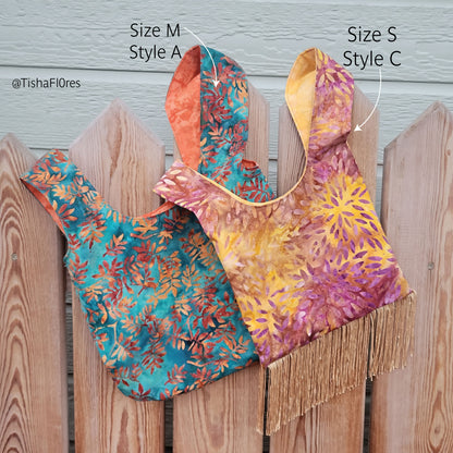 Two patterned fabric shopping bags made from a sewing pattern displayed on a wooden fence, labeled with arrows and text as Size M Style A and Size S Style C. One bag has a teal, orange, and gold leaf pattern, and the other has a yellow, orange, and purple floral pattern with fringe at the bottom.