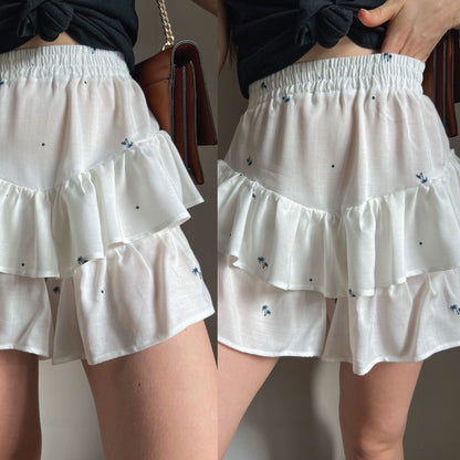 A close-up split view of a person wearing a white tiered ruffle mini skirt with small blue floral embroidery and tiny black dot accents. The skirt has an elastic waistband and is styled with a black top and a brown handbag.
