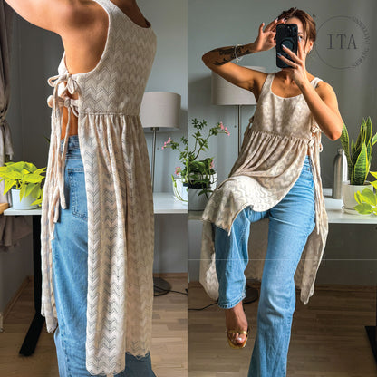 A woman modeling a sleeveless cream knit cardigan with a subtle chevron pattern and tie-back detail, paired with light blue jeans in a mirror selfie inside a home setting.