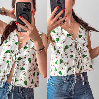 A split-screen mirror selfie showing a woman wearing a white sleeveless tie-front top with an avocado print and blue jeans. The top features a repeating pattern of green avocados and red/pink seed details on a white background.