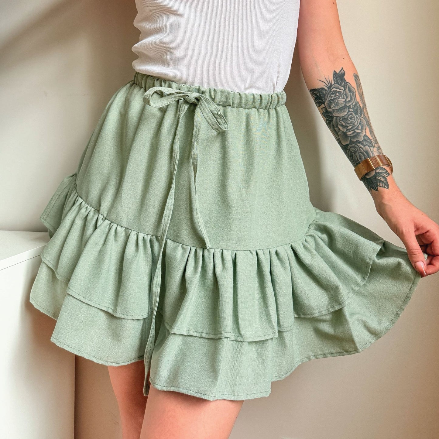 A person wearing a light sage green tiered ruffle mini skirt with a drawstring waist, paired with a white ribbed top. The photo is cropped from the chest down, showing the model's tattooed arm holding out the side of the skirt.