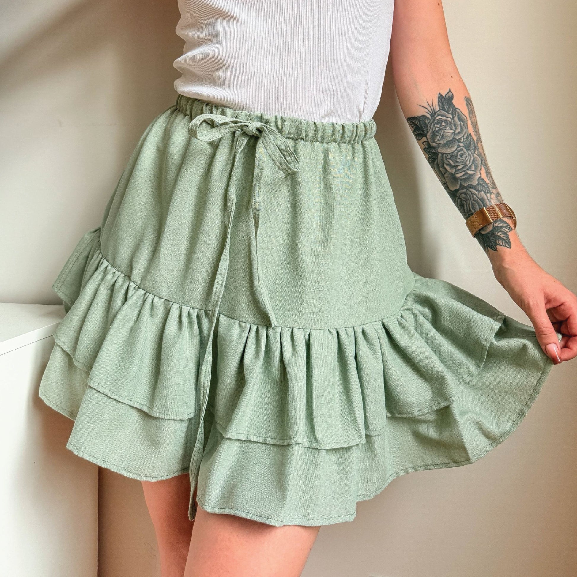 A person wearing a light sage green tiered ruffle mini skirt with a drawstring waist, paired with a white ribbed top. The photo is cropped from the chest down, showing the model's tattooed arm holding out the side of the skirt.