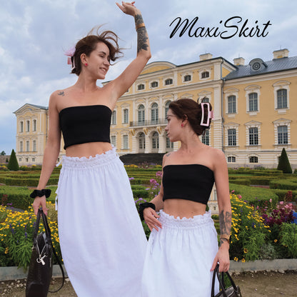 Two women standing outdoors in front of a large yellow historic building, wearing matching black bandeau crop tops and white maxi skirts. The word 'MaxiSkirt' appears in script at the top right.