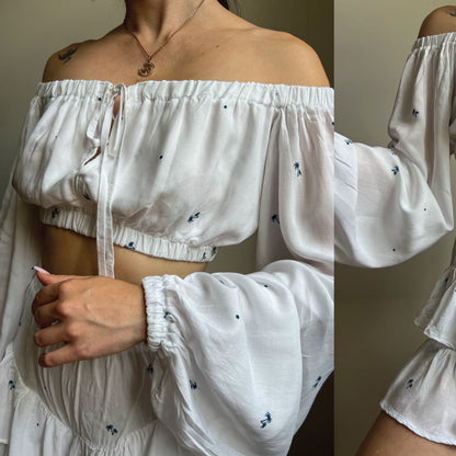 White off-the-shoulder crop top with long balloon sleeves and small blue embroidered details, shown on a model in a split-view photo.