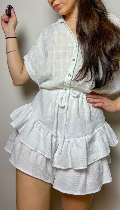 Woman wearing finished Velora shorts sewing pattern in white fabric with ruffled layers and tie waist, shown against a neutral background.
